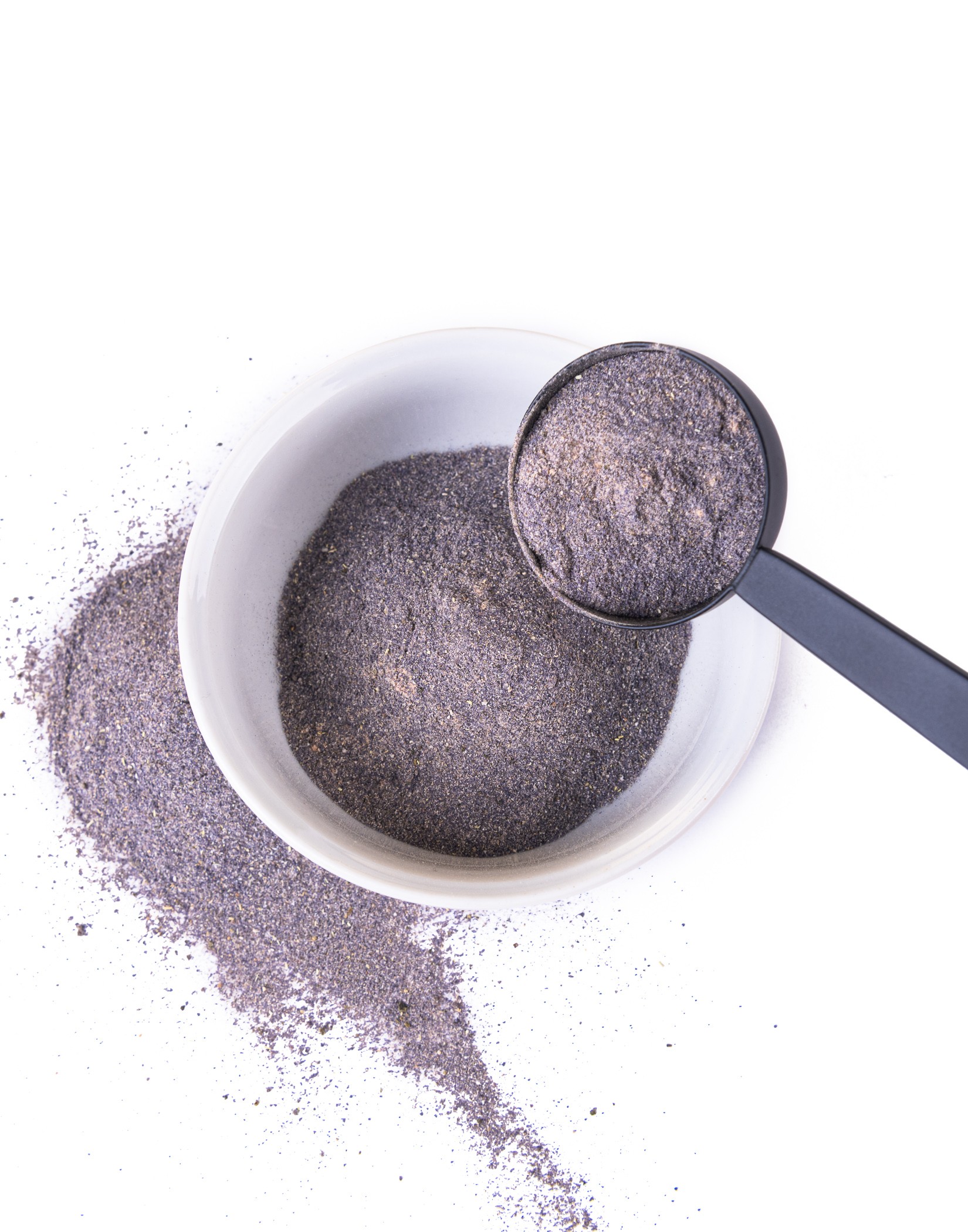A white bowl with dark purple powder, next to it scattered powder and a black spoon with a portion of the powder.