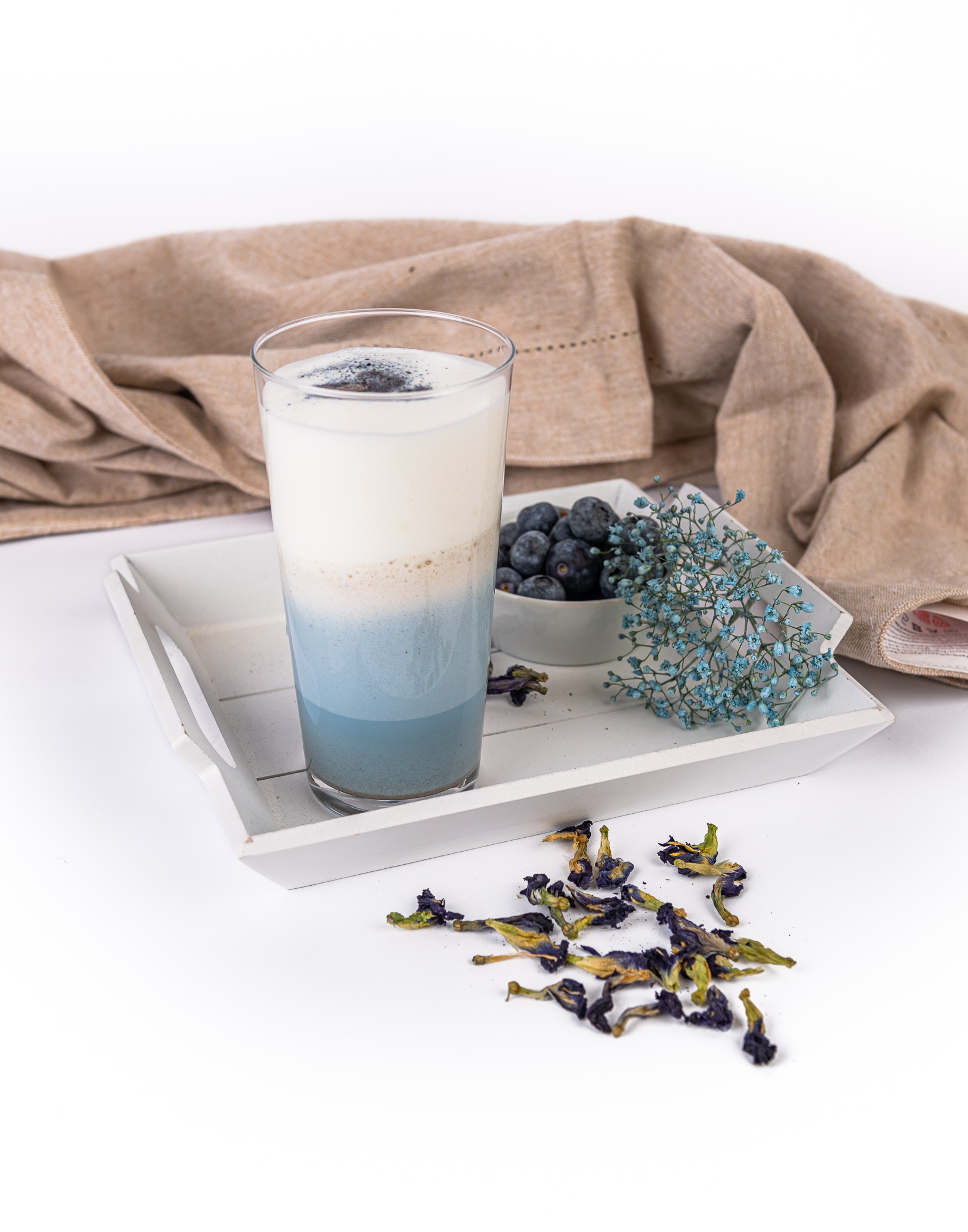 A glass of blue drink with foam and sprinkles, next to berries, light blue flowers and dried on a white tray.