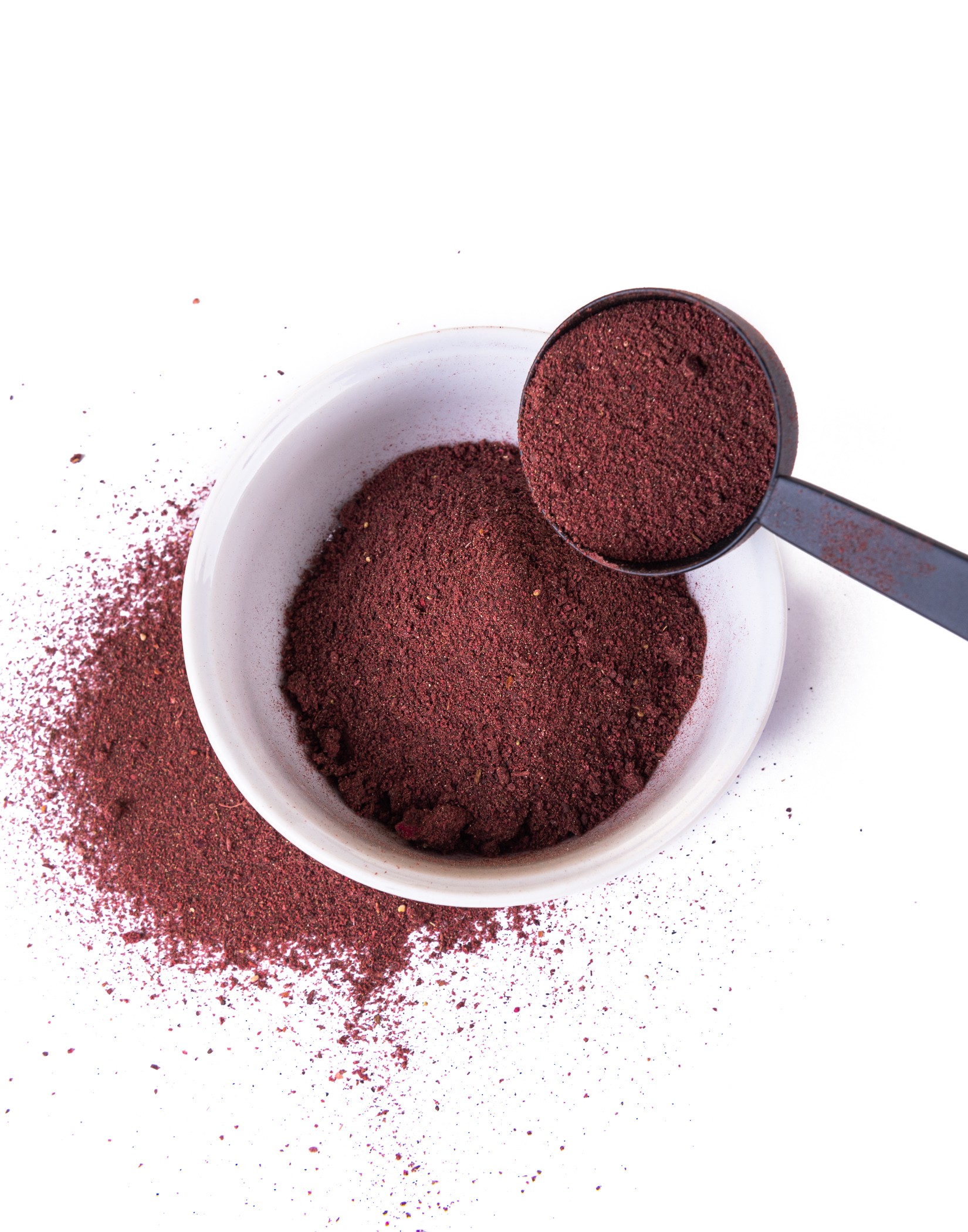 Biała miska z czerwonofioletowym proszkiem, obok rozsypany proszek i czarna łyżka z porcją proszku.