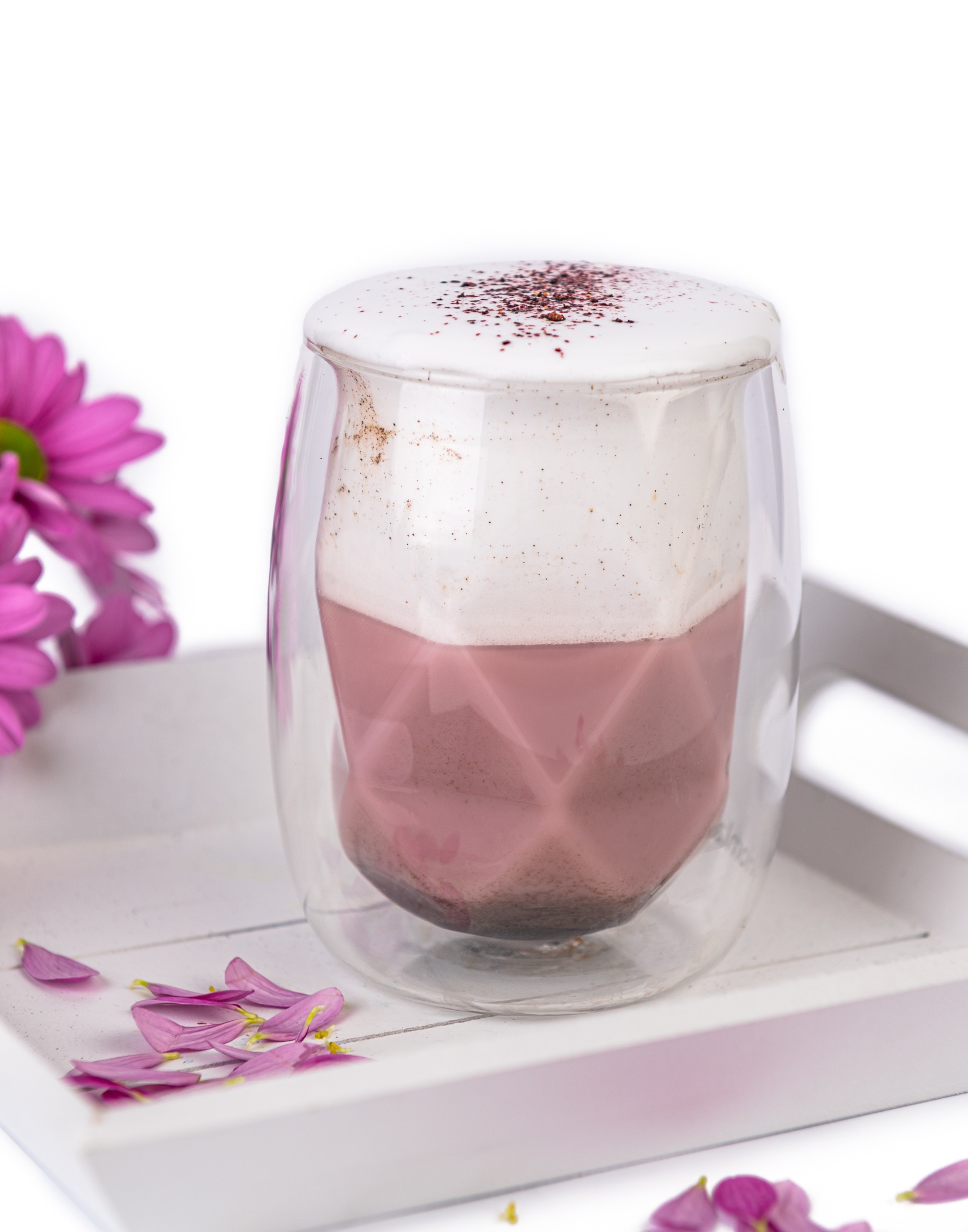 Szklanka z pianką i różowym napojem na tacy, wokół płatki fioletowych kwiatów i kwiat z lewej strony.
