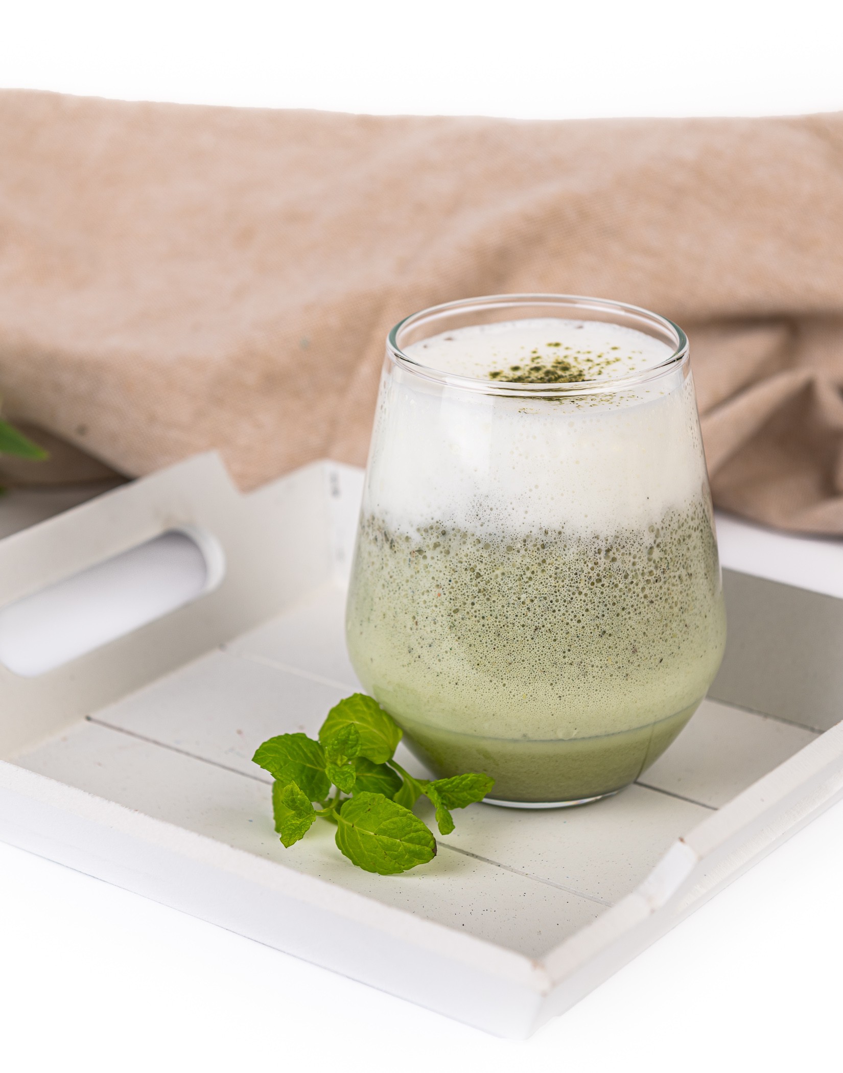 Szklanka matcha latte z mleczną pianką na tacy, obok zielona gałązka mięty.
