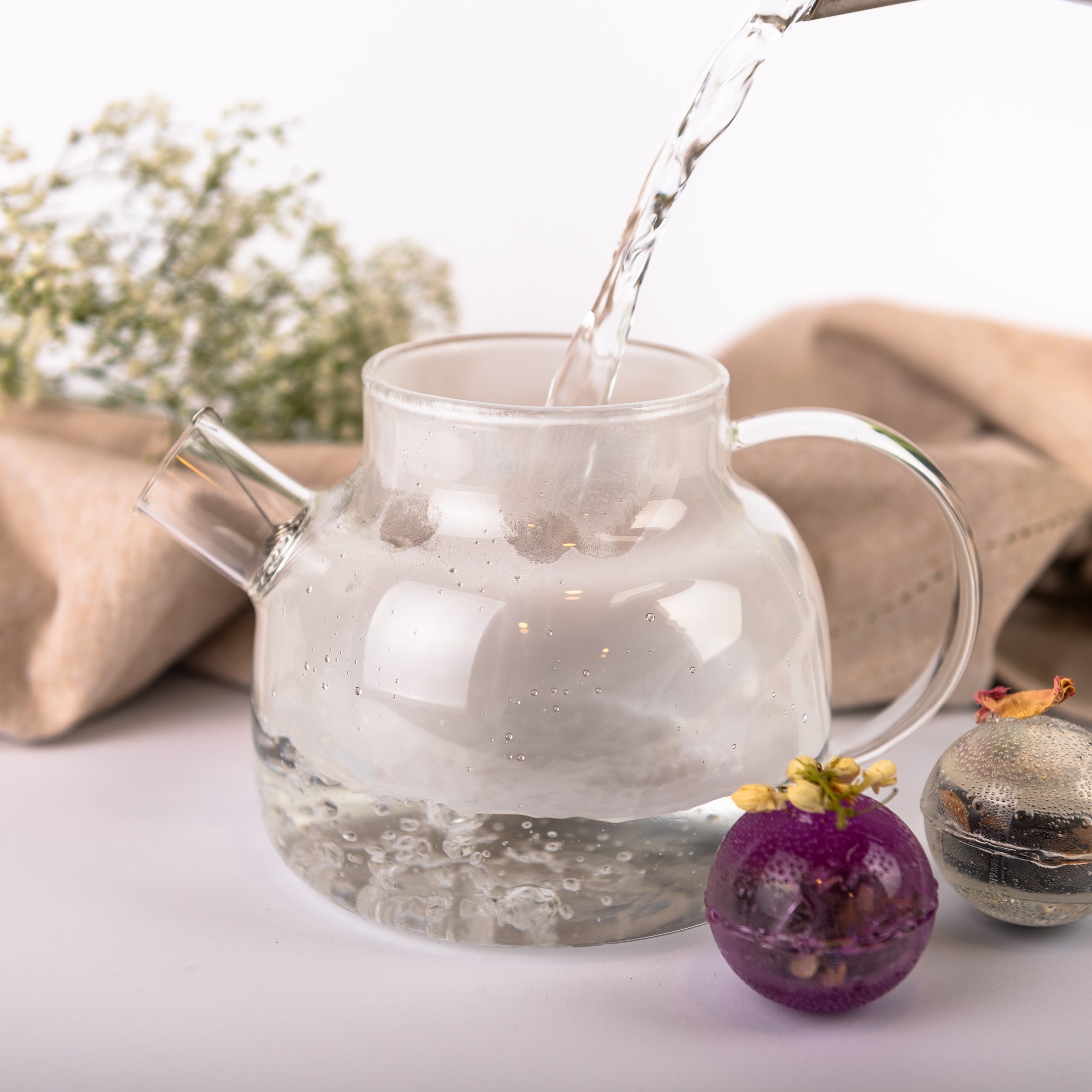 Hot water is poured into a glass pitcher. Next to it lie two colorful flower balls for brewing tea.