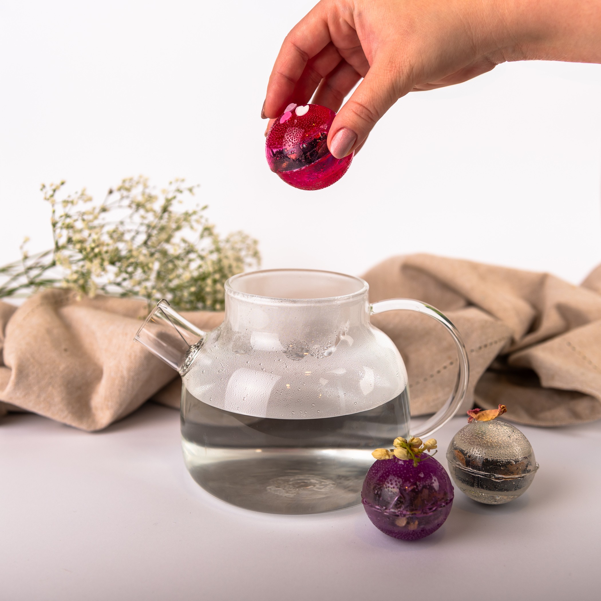 A glass teapot with hot water and two colorful flower tea balls; one ball held above the teapot by a hand.