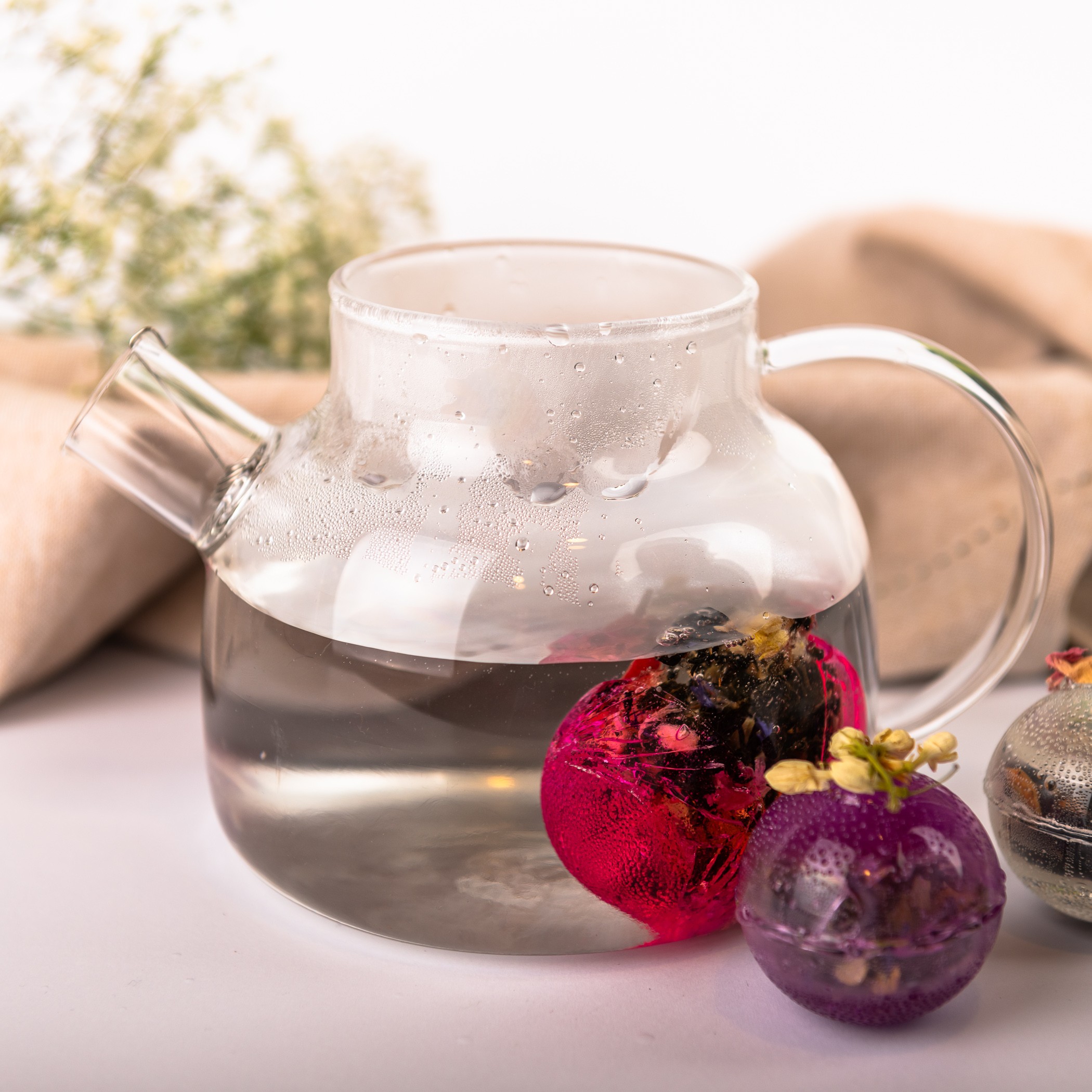 A glass teapot with hot water and two colorful flower balls for brewing tea; one ball is already brewing, the other lies next to the teapot.