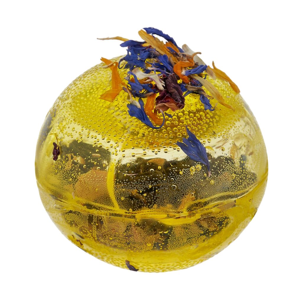 Kula do parzenia herbaty w intensywnie żółtym kolorze, z ziołowo-kwiatową posypką na szczycie.