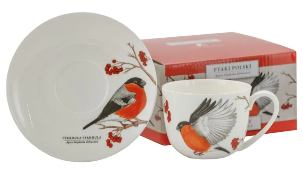 Filiżanka ze spodkiem ozdobiona ilustracją gila z czerwonym brzuszkiem oraz gałązkami jarzębiny, w tle czerwone pudełko z tym samym motywem.