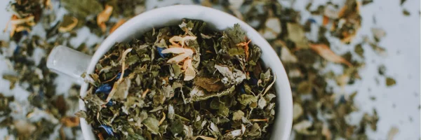 Biała filiżanka wypełniona mieszanką suszonych liści i kwiatów, wokół niej rozsypane zioła.