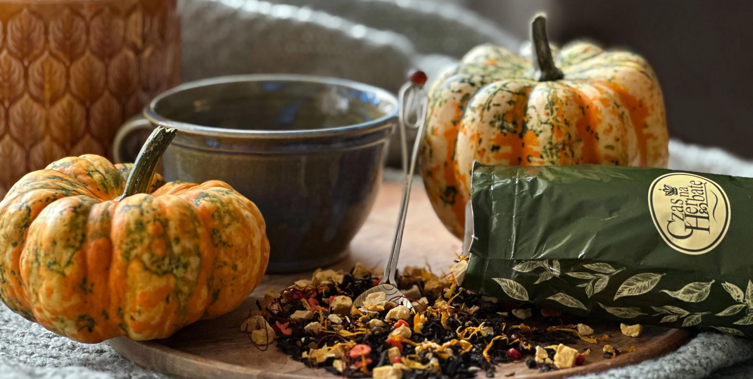 Filiżanka oraz mieszanka herbaty owocowej wysypana na drewnianej desce, z dwoma małymi dyniami i opakowaniem herbaty jesiennej marki Czas na Herbatę.