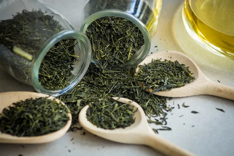 Rozsypane liście zielonej herbaty wysypujące się ze słoików, obok drewniane łyżki i filiżanka naparu.