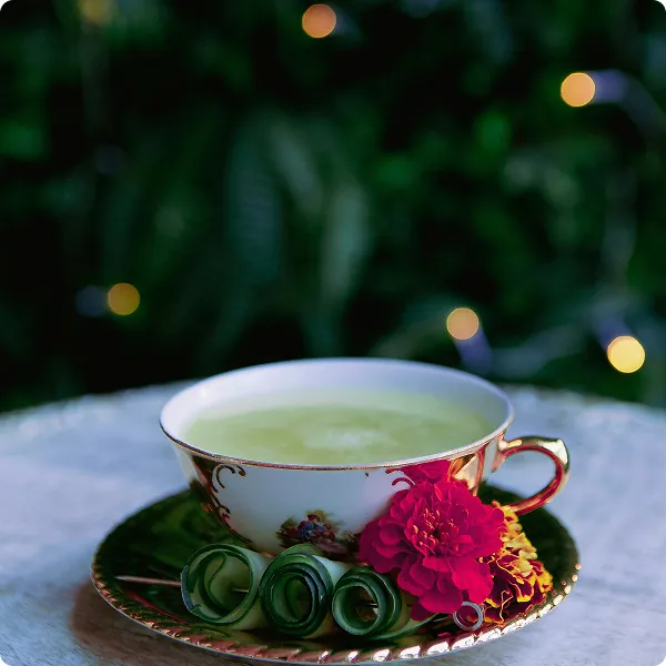 Porcelanowa filiżanka z jasnozielonym naparem na ozdobnym spodku, obok dekoracyjna kwiatowa ozdoba; w tle widoczne rozmyte światła i liście