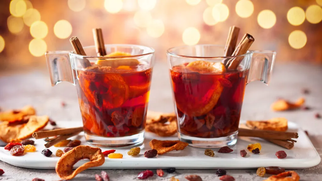 Dwa szklane kubki z kompotem z suszu z owocami i laskami cynamonu w środku na tle światełek