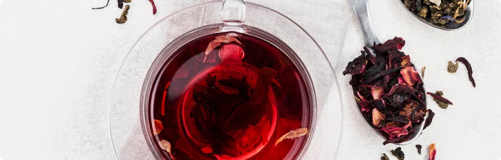 Szklany kubek z czerwoną herbatą widziany z góry, obok łyżka z suszonymi płatkami kwiatami